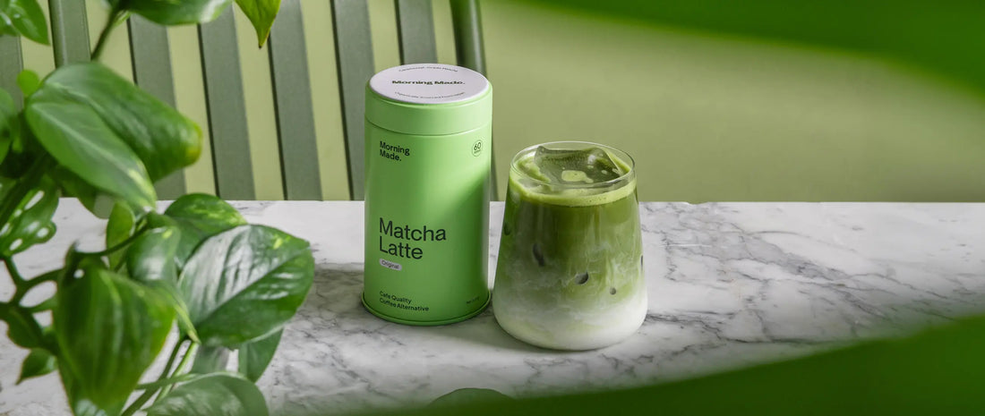 Image of matcha latte tin with glass of matcha sitting beside it. Both sitting on a marble benchtop, with a leafy plant to the side.
